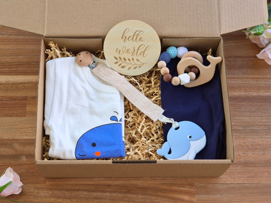 A baby gift set in a cardboard box filled with shredded paper, featuring a white t-shirt with a blue whale graphic, a navy leggings, a wooden sign reading 'hello world', a wooden bead teether ring, and a muslin pacifier with silicone whale.
