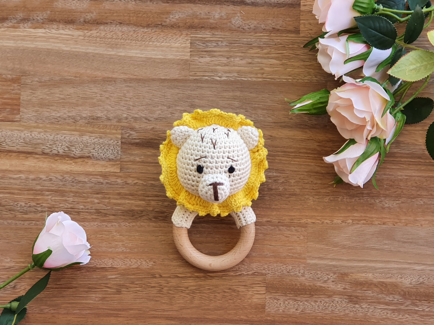 A crocheted lion baby Rattle with a yellow mane, cream face, and a wooden ring handle