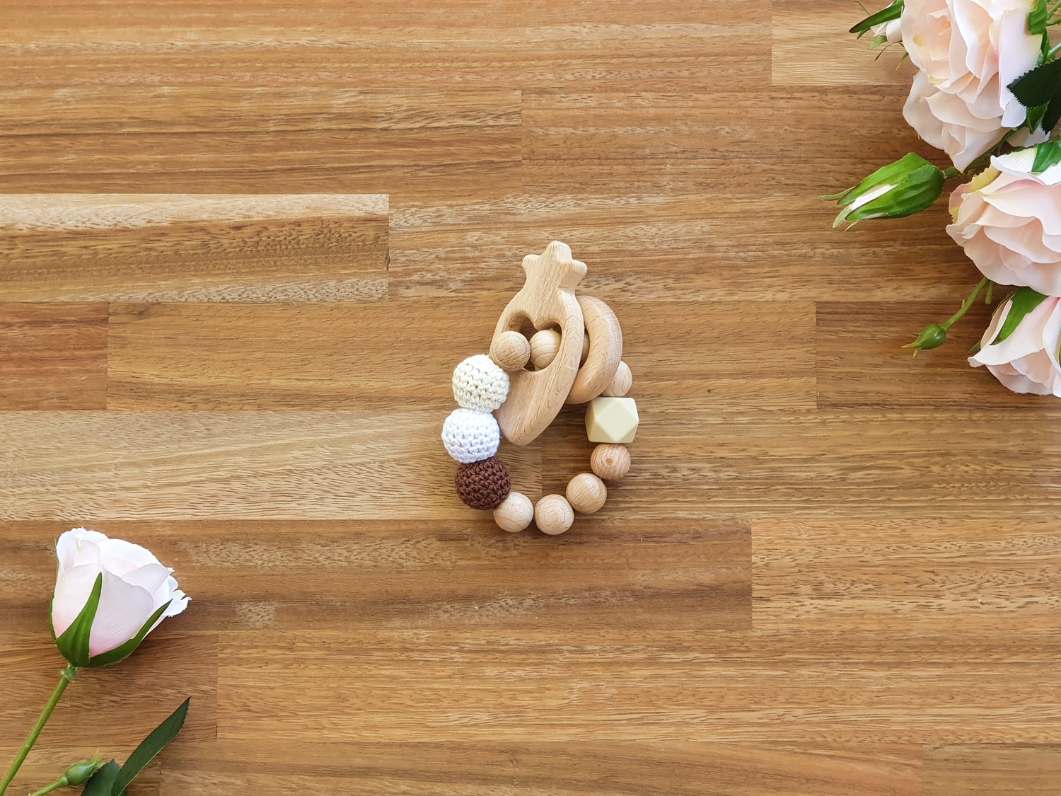A wooden beaded baby teether ring with knitted beads in natural wood, white, and dark brown, styled on a light brown wooden surface with soft pink roses in the corners.