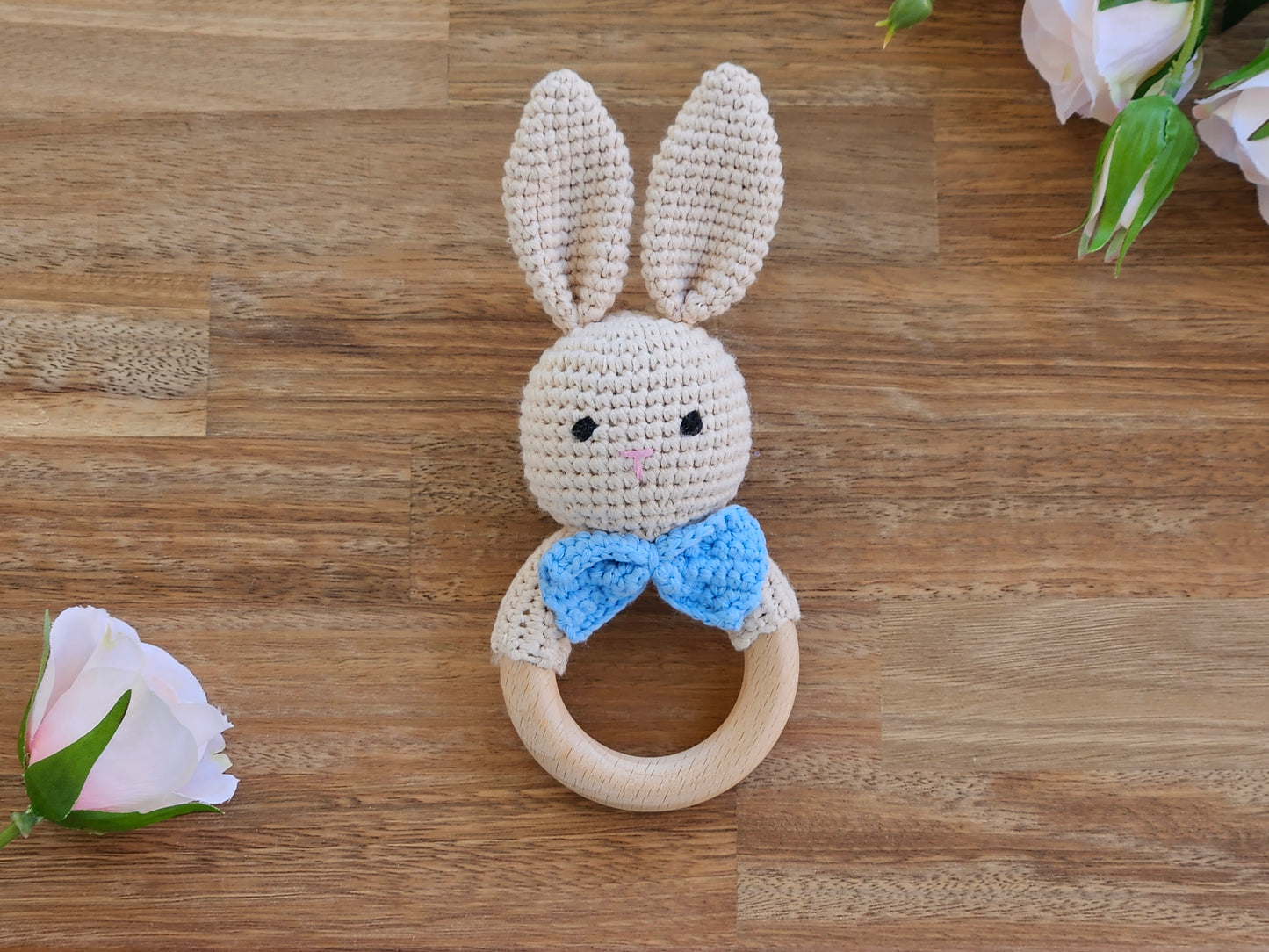 A crocheted bunny teething ring rattle with long cream-colored ears, a blue bow tie, and a natural wooden ring, 