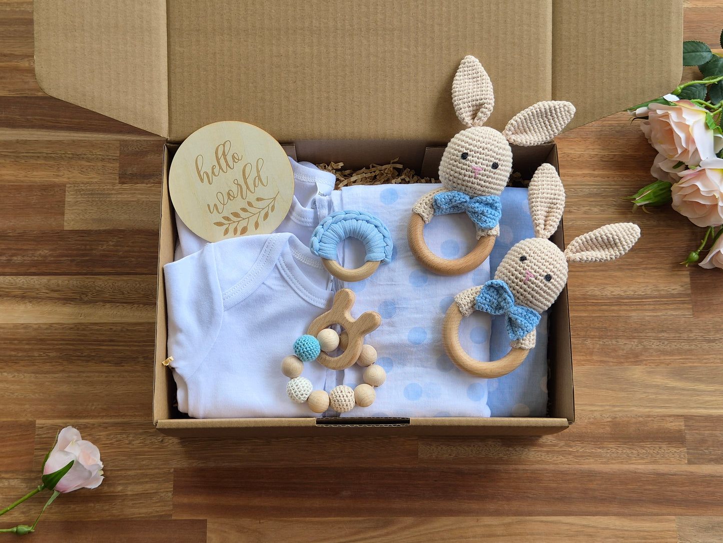 A twin baby gift box set , featuring  2 white baby bodysuits, two crocheted bunny teething rattles with blue bows, a wooden bead teether, 2 cotton wraps, a round wooden Hello world disc,  packaged in a gift box.