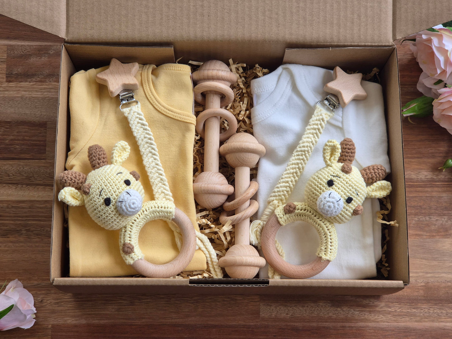 A baby gift box arranged in an open cardboard box, featuring a yellow and white baby romper set with wooden and knitted giraffe teething rings and a wooden barbell rattle.