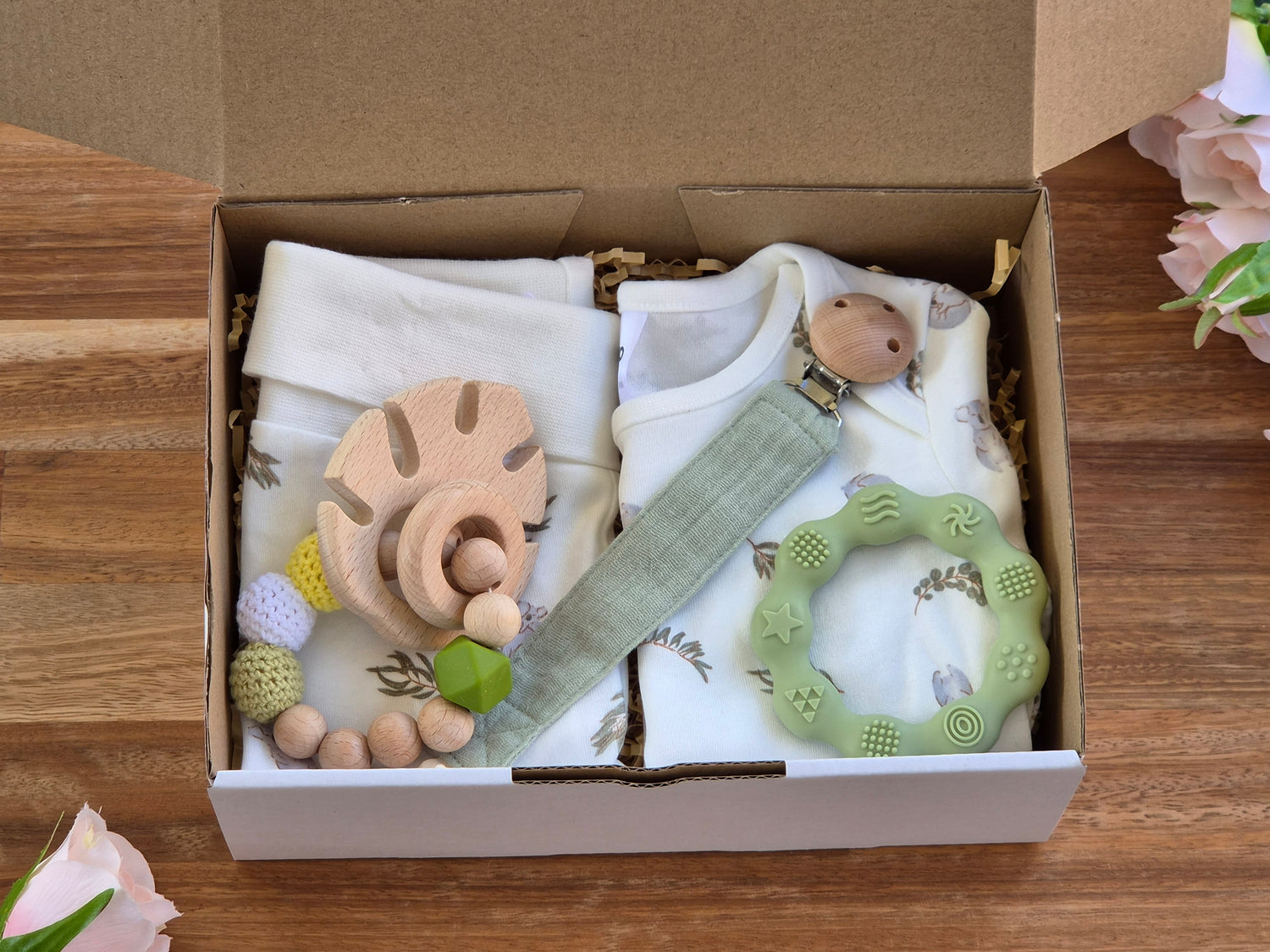 A baby gift set inside an open kraft box, featuring cream coloured baby clothes with a subtle leaf and koala pattern, a light green muslin pacifier clip, a wooden and beaded leaf teether, and a green silicone teether ring.