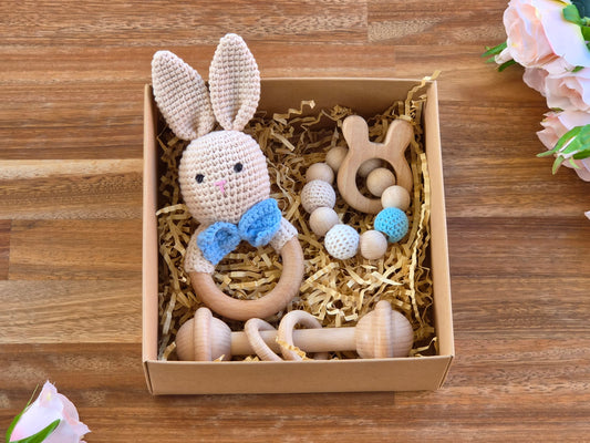 A baby gift set in a brown cardboard box filled with shredded paper, featuring a crocheted bunny rattle with a blue bow, wooden teething toy bar and beaded wooden teether ring displayed on a wooden tabletop with pink flowers in the corners.