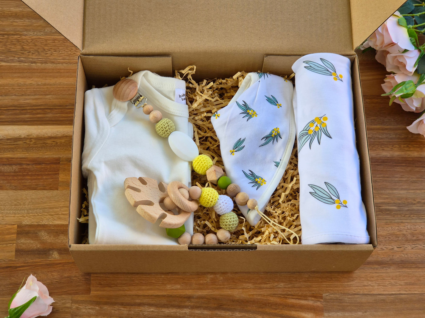 A baby gift set arranged inside an open brown cardboard box filled with shredded paper. The set includes a white baby bodysuit, a white cotton swaddle blanket with a green and yellow wattle print, a matching baby bib and a wooden bead teether ring and pacifier clip.
