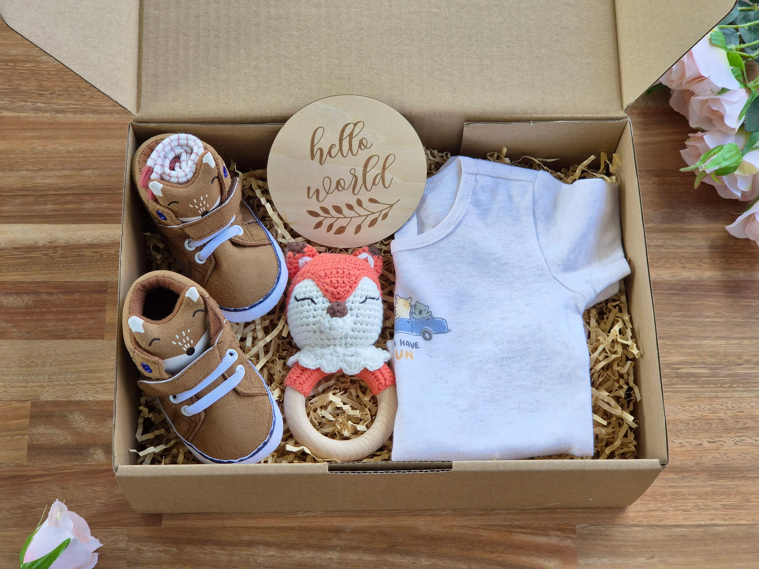 A baby gift box set containing a white short-sleeve bodysuit, a pair of brown baby Fox face design boots, a pair of socks, crocheted fox teether, and a round wooden sign that reads 'hello world', arranged in a cardboard box with shredded paper on a wooden table.
