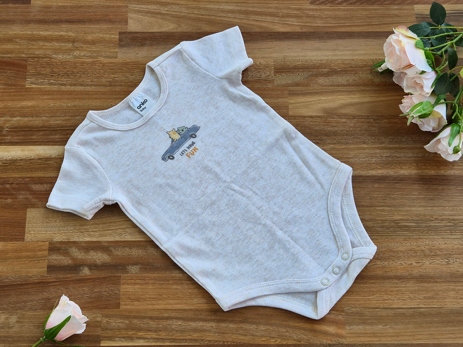 A light heather grey short-sleeve baby bodysuit with a small graphic of a blue car and the word 'FUN' in yellow, laid flat on a wooden surface with pink roses in the corners.
