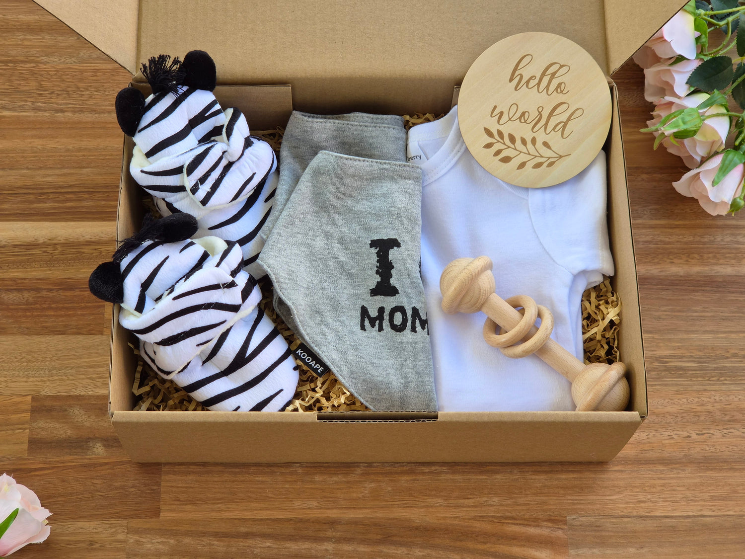 A baby gift box filled with a white onesie, 2 grey bandana bibs with 'I' love MOM' and 'I love DAD' text, a zebra-print slippers and a wooden teething toy and a wooden disc with text Hello World.