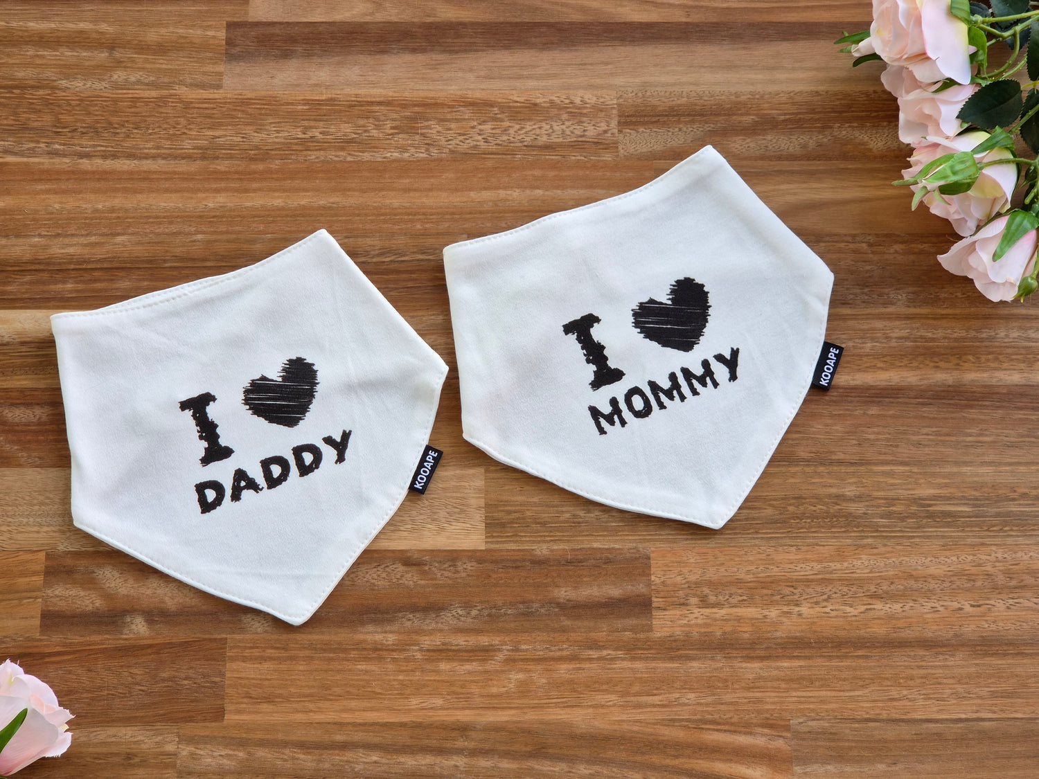 Two white triangular bandana bibs laid flat on a wooden surface. One bib reads 'I [heart] DADDY' and the other reads 'I [heart] MOMMY' in black text with a black heart.