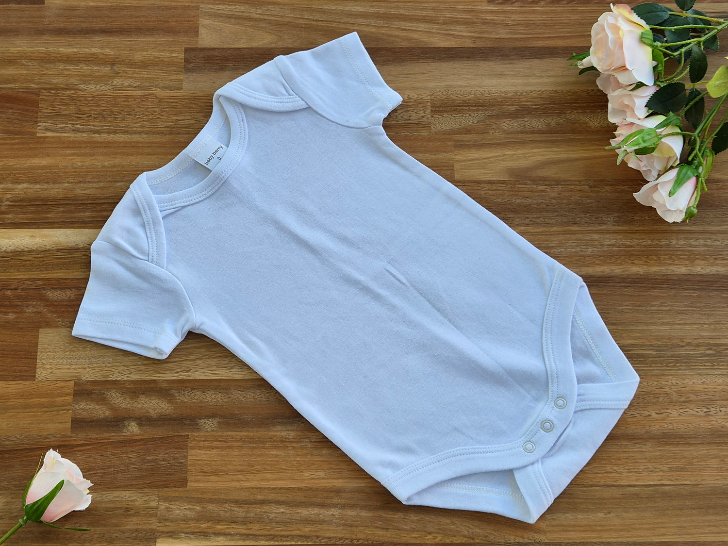 A white short-sleeve baby bodysuit laid flat on a wooden surface, styled with pale pink roses in the corners.