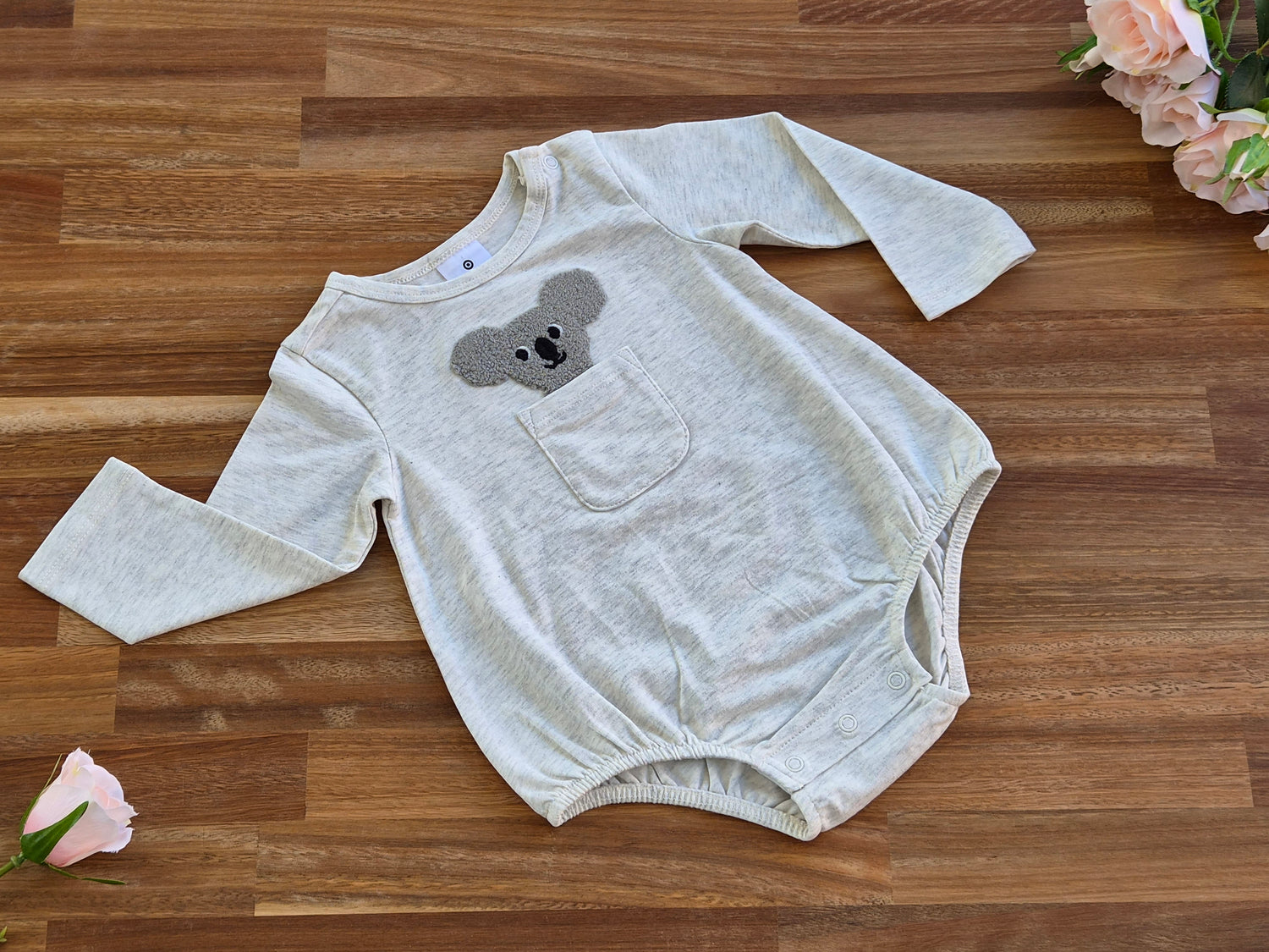 A light heather grey long-sleeve baby bodysuit with a grey koala face patch on the chest pocket,.