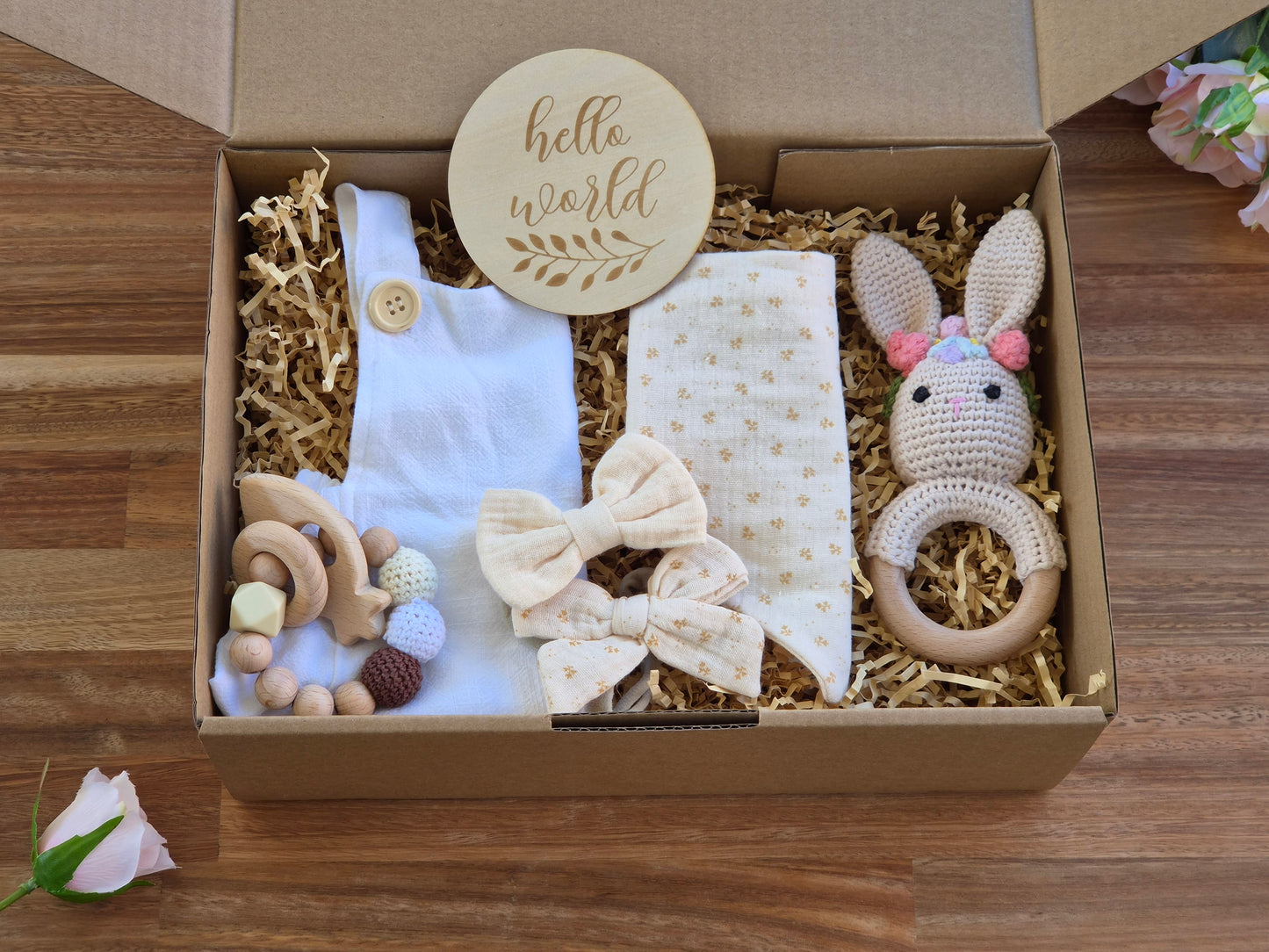 A baby gift set in an open cardboard box filled with shredded paper, featuring a bib brace white onesie, a cream bandana bib with 2 matching hair bows, a knitted bunny rattle, wooden beaded teething ring, A round wooden disc reading 'hello world' is placed inside the box on a wood surface.