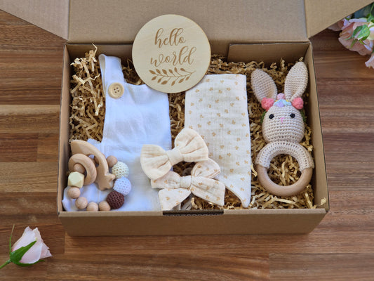 A baby gift set in an open cardboard box filled with shredded paper, featuring a bib brace white onesie, a cream bandana bib with 2 matching hair bows, a knitted bunny rattle, wooden beaded teething ring, A round wooden disc reading 'hello world' is placed inside the box on a wood surface.